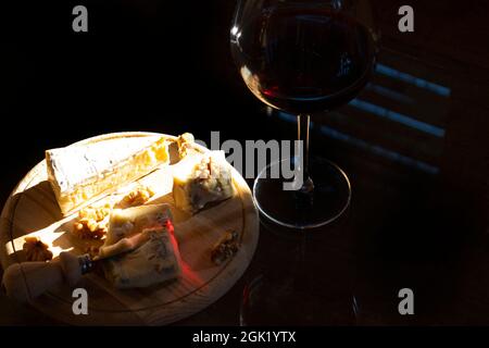 soft cheeses Brie and gorgonzola on wooden cutting board, walnuts,  red wine in the goblet i, n the dark Stock Photo