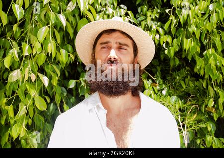 young man with beard and long hair wears hat and white shirt surrounded by greenery Stock Photo