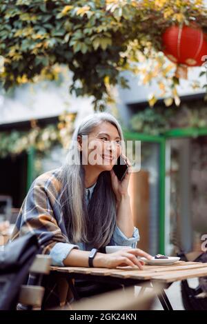 Positive mature Asian woman talks on cellphone at small table in outdoors cafe Stock Photo