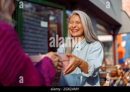 Cheerful mature Asian woman with friend hold gift box sitting at small table in cafe Stock Photo