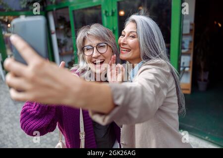 Positive senior Asian lady with grey haired friend take selfie on modern city street Stock Photo