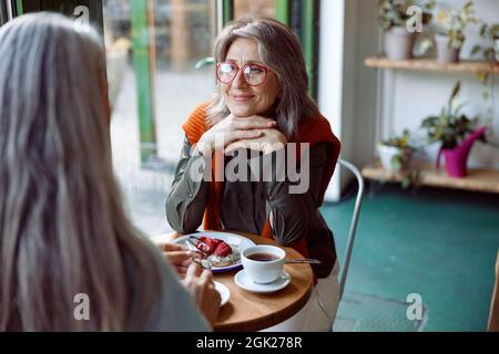 Grey haired woman with glasses looks at friend sitting at small table in cozy cafe Stock Photo