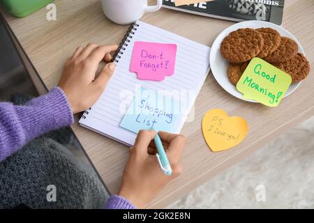 Woman writing text SHOPPING LIST on sticky note at home Stock Photo