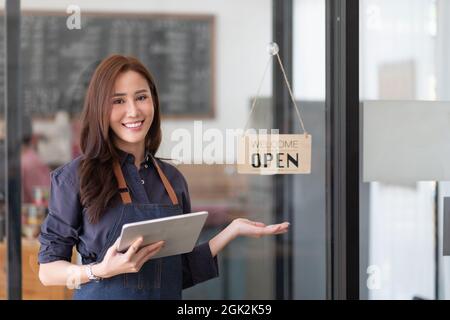 Portrait of a smiling Asian entrepreneur standing behind her cafe counter with open sign board Stock Photo
