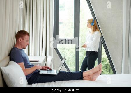 Pretty melancholic young woman listening to good music in headphones and looking through window, her husband in working on laptop Stock Photo