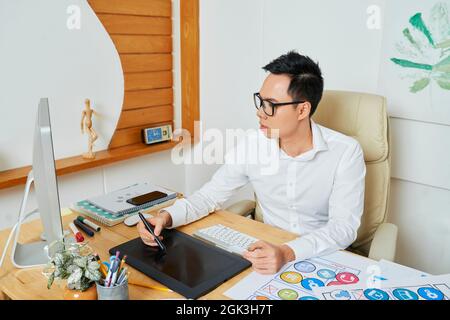 Serious young Asian graphic designer in glasses drawing logotype or set of icons for mobile application Stock Photo