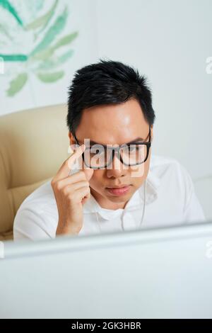Frowning serious graphic designer adjusting glasses when reading project technical requirements on computer screen Stock Photo