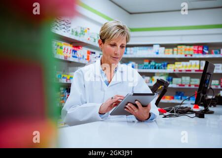 Caucasian female pharmacist scrolling on digital tablet while standing in pharmacy drugstore Stock Photo