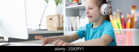 Little girl in headphones in front of computer is typing on keyboard Stock Photo