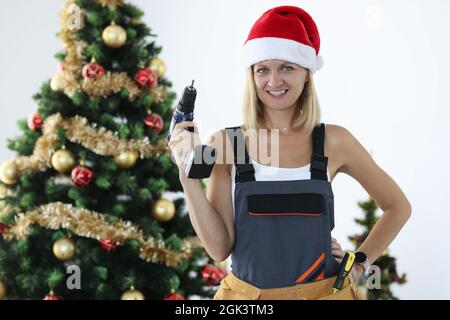 Smiling woman repairman in uniform and santa claus hat holding drill in hands near christmas tree Stock Photo