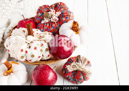 Cozy pumpkins. Autumn decoration with handmade colorful fabric pumpkins, red apples and white knitted sweater on white wooden background. Fall vibes. Stock Photo