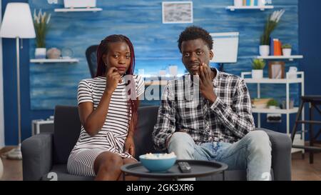 POV of african american couple being shocked watching TV drama movie in living room. Black married people looking at camera while sitting on couch at home. Partners relaxing together Stock Photo