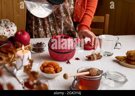 Cozy autumn days. Woman wearing orange sweater pouring hot water for brew tea in red teapot on the table with linen tablecloth. Aromatic mood. Fall vi Stock Photo