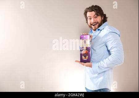 Riga,Latvia- August 4, 2021:  Happy bearded man holds in his hands a box of Courvoisier VSOP Cognac  on a light gray background. Stock Photo