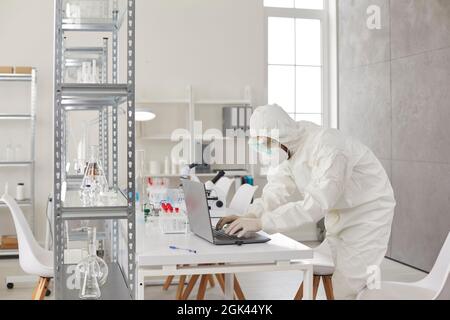 Scientist in PPE coveralls, goggles, mask and gloves working on laptop in medical laboratory Stock Photo