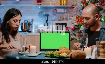 Couple eating festive meal watching horizontal green screen on laptop. Man and woman enjoying christmas eve dinner, having chroma key with mockup background and isolated template Stock Photo