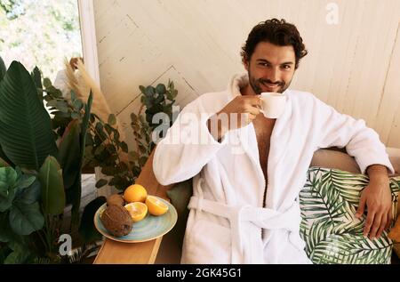 Handsome man drinking wellness tea while relaxing at exotic spa resort, wearing a white bathrobe and sitting on armchair Stock Photo