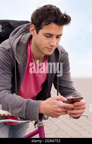 vertical photo of a handsome young man leaning on a bike using a mobile phone, concept of communication and ecological urban transportation Stock Photo