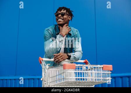 afro shopping with a shopping cart, the guy is shopping near the mall, a young black student on the background of a blue building Stock Photo