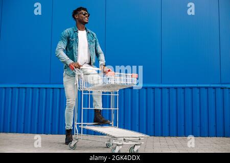 afro shopping with a shopping cart, the guy is shopping near the mall, a young black student on the background of a blue building Stock Photo