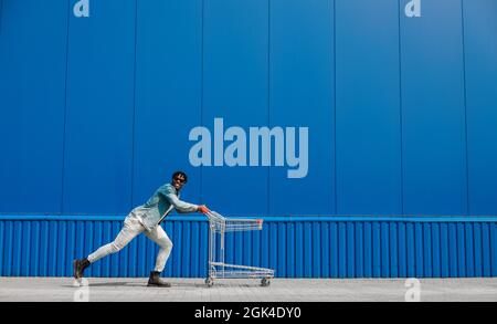 black afro guy shopping with a shopping cart, guy having fun and jumping, runs shopping near the mall, young black student on background of a blue bui Stock Photo