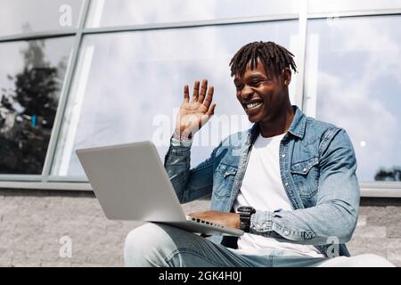 black african american man typing on a laptop on the street in front of a glass building sitting, talking skype, high-speed mobile internet, 5g, 4g, w Stock Photo