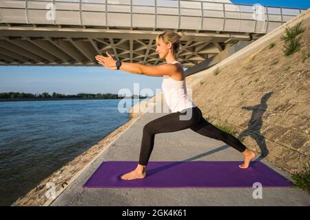 Young woman is exercising outdoor. She is practicing yoga on sunny day. Virabhadrasana / Variation of Warrior pose. Stock Photo