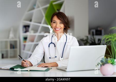 Smiling female healthcare worker doing some paperwork and using laptop while working at doctor's office. Stock Photo