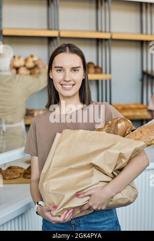 Portrait of happy young female customer standing with paperbag full of loaves of bread in bakery store Stock Photo