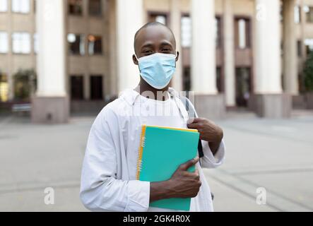 African american male student in face mask standing outdoors with notebooks, posing to camera near university building Stock Photo