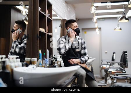 Young man haidresser using smartphone in barber shop, coronavirus and new normal concept. Stock Photo