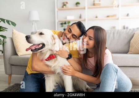 Portrait of happy multiracial couple scratching their pet dog, sitting on floor at home Stock Photo