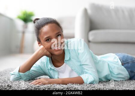 Smiling young pretty black girl student in casual looks at camera and rests, lies on floor in living room interior Stock Photo