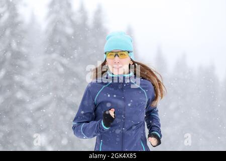 Young slim woman running on the road in the winter snowy morning. Close up portrait of jogging fitness woman. Stock Photo