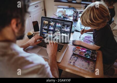 Unrecognizable father with two small children working indoors at home, painting pictures and home office concept. Stock Photo
