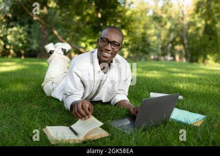 Happy african american student guy lying on grass at park, preparing for classes, using laptop and book outdoors Stock Photo