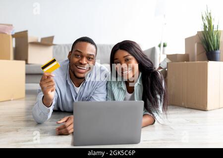 Loving black spouses buying house decor online, using laptop and credit card, lying on floor among carton boxes Stock Photo