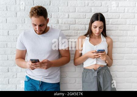 Two friends or lovers standing front of awall and looking to their mobile phones. Technology addiction concept. High quality photo Stock Photo