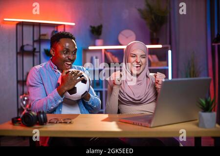 Overjoyed muslim woman and african man gesturing and screaming while watching football match on laptop. Young mixed race couple celebrating win of favorite team. Stock Photo