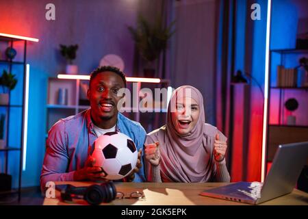 Portrait of african man and muslim woman emotionally cheering favorite team during soccer game. Young couple using modern laptop for watching match at home. Stock Photo