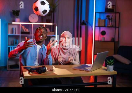 African american man throwing up soccer ball while muslim woman in hijab looking at camera with worried facial expression. Young couple spending evening time at home for watching football game. Stock Photo