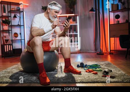 Caucasian aged man in sportswear and headband surfing internet on smartphone while sitting on balance ball. Concept of retirement, active lifestyles and modern gadgets. Stock Photo