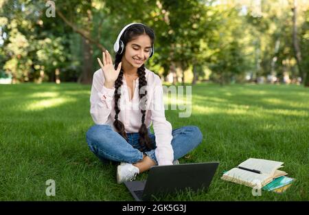 Remote learning. Excited indian female student using laptop, having online lesson and waving to webcam, sitting outdoors Stock Photo