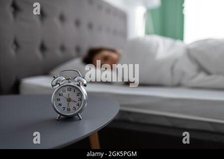 Alarm clock on bedside table and blurred view of young lady lying in bed, covered with blanket, unwilling to wake up Stock Photo