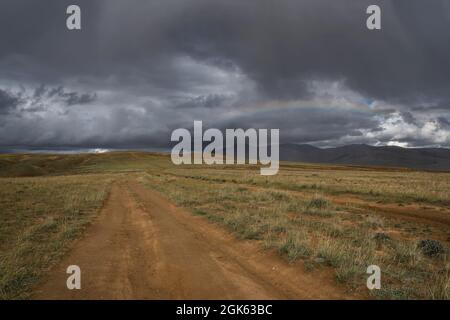 Amazing rainbow on a dark stormy sky with thunderclouds and rain over the mountains and a dirt road in the steppe. Altai, Russia Stock Photo