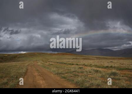 Amazing rainbow on a dark stormy sky with thunderclouds and rain over the mountains and a dirt road in the steppe. Altai, Russia Stock Photo