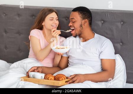 Affectionate multiracial couple having breakfast in bed, millennial lady feeding her boyfriend with cereal at home Stock Photo