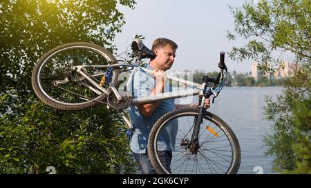 A frustrated man carries a broken bicycle on his shoulders by the river in the park. Stock Photo