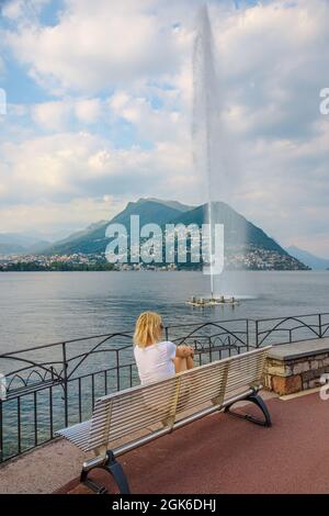 woman looking water jet of Paradiso fountain of Lugano city in Lugano Lake of Switzerland. Bench by the waterfront of Lugano Lake with Monte San Stock Photo