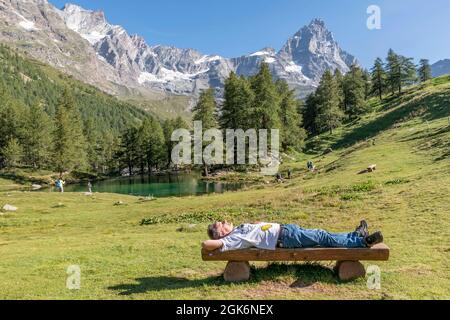 A white man lying in the sun on a wooden bench enjoying the tranquility of Lago Blu, Aosta Valley, Italy Stock Photo